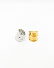 Coppia di anelli Odine in acciaio inossidabile colo argento e oro. L'anello argento è rappresentato in 3/4 e fa vedere il retro regolabile, quello oro è fotografato frontalmente e evidenzia il design a onde con una fascia più spessa al centro.
