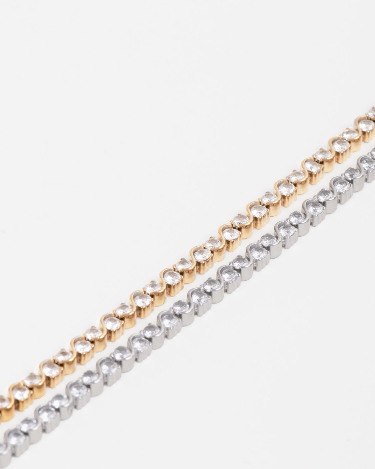 Coppia di bracciali Monica, in acciaio inossidabile e strass, modello tennis con motivo ad S. Sono rappresentati su sfondo bianco: il bracciale di destra è di color oro mentre quello di sinistra è argento.
