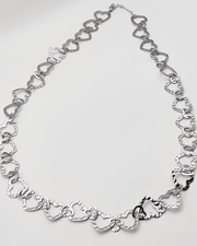 Collana Teresa in color silver. E' formata da tanti cuori in acciaio martellato intrecciati tra loro. E' ripresa su uno sfondo grigio chiaro.