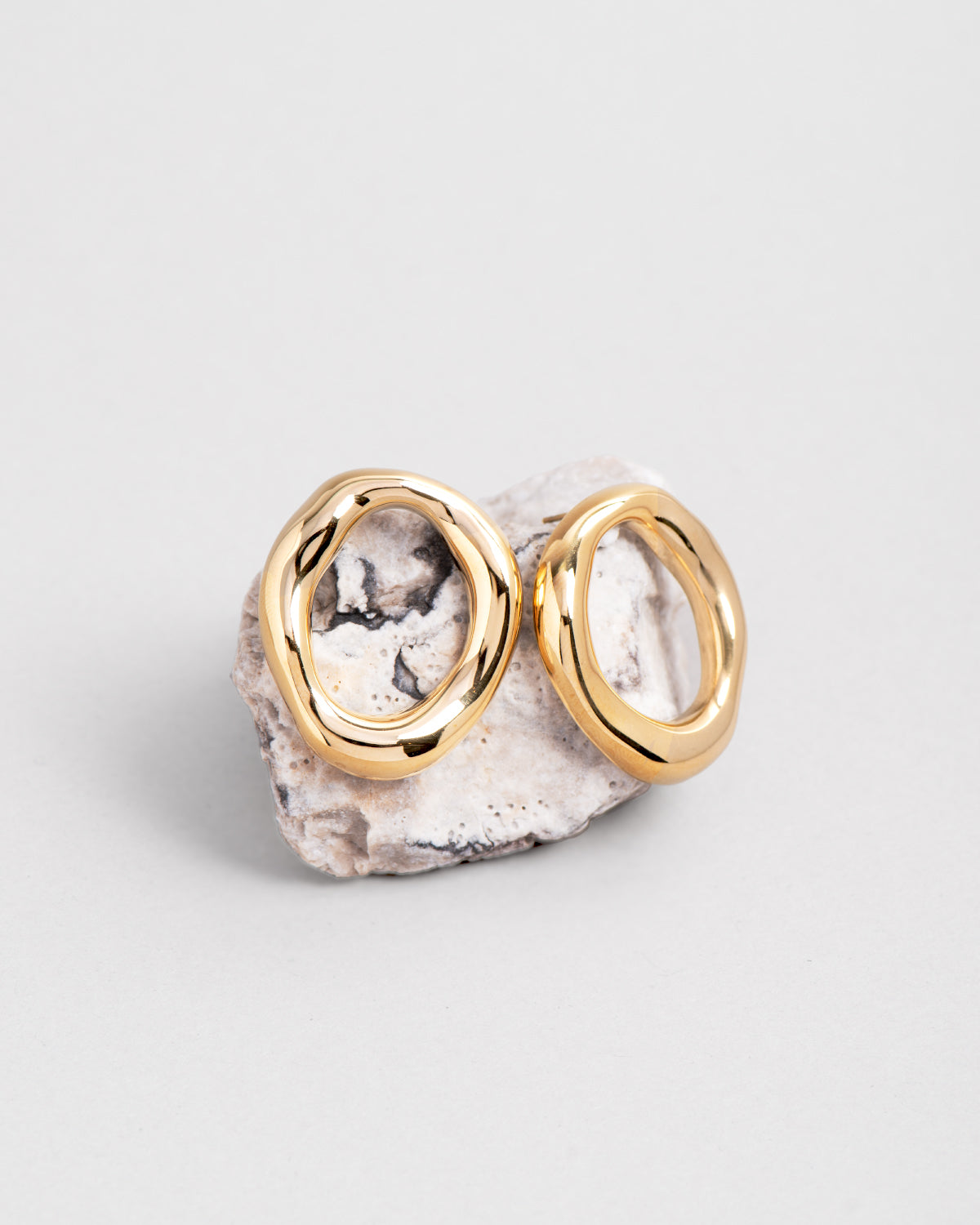 Gli orecchini  in acciaio inossidabile Denise sono posizionati su una roccia dai toni del bianco e del nero. Hanno una forma ovale irregolare di colore oro lucido.