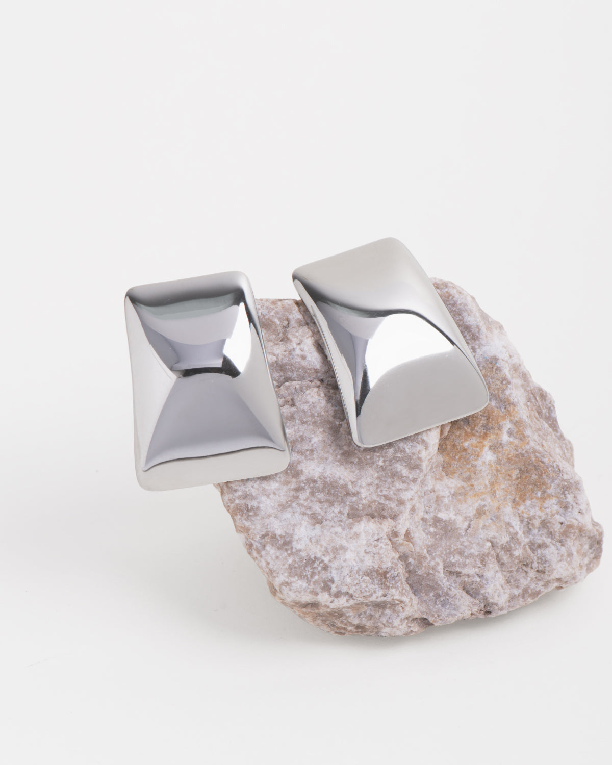 Gli orecchini Elia in acciaio inossidabile color argento sono posizionati su una roccia dai toni bianchi e aranciati su sondo bianco. Hanno una forma rettangolare con una bombatura centrale e finish lucido. Sono più stretti nella parte superiore e più larghi in quella inferiore. 
