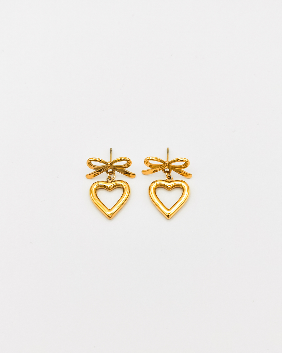 Orecchini Emma in acciaio inossidabile color oro. Sono formati da un fiocchetto che si fissa al lobo con una chiusura a perno, e un pendente a forma di cuore traforato.