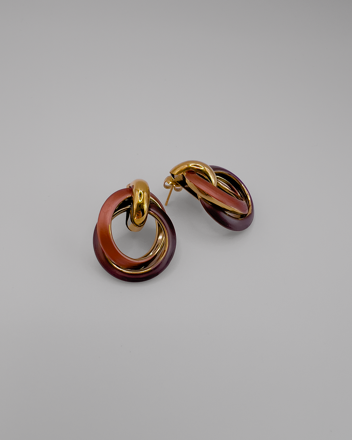 Orecchini Ivana in acciaio inossidabile color oro e smalto in due tonalità di marrone. Sono formati da due cerchi intrecciati legati da un semicerchio dal quale parte il perno per indssarli. L'orecchino di sinistra è frontale mentre quello di destra è di lato.