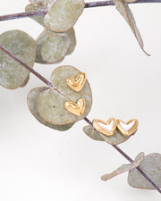 Doppia coppia di orecchini Matilde in acciaio inossidabile color oro in due versioni: lucidi o con centro in madreperla, entrambi a forma di cuore asimmetrico. Sono appesi a delle foglie di eucalipto su uno sfondo bianco.