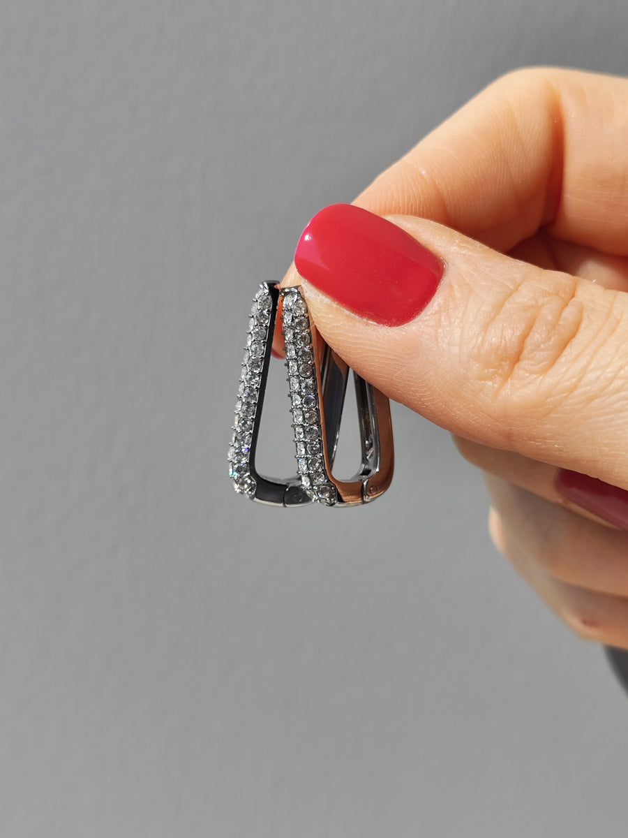 Orecchini Lorena in acciaio inossidabile color argento con tripla fila di strass.Sono fotografati lateralmente sorretti da una mano femminile su sfondo grigio.