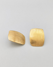 Gli orecchini Marika in acciaio inossidabile anallergico color oro sono rappresentati frontalmente su uno sfondo grigio chiaro. Hanno una forma rettangolare con delle righe verticali leggermente ondulate. L'orecchio di sinistra è ripreso lateralmente mentre quello di destra frontale.