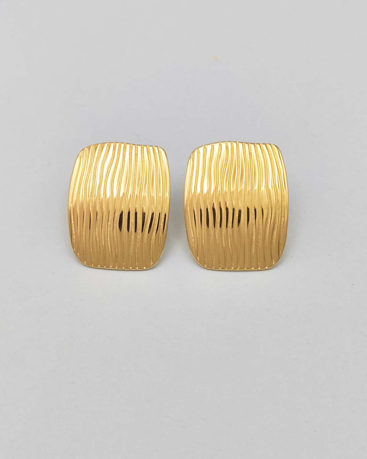Gli orecchini Marika in acciaio inossidabile anallergico color oro sono rappresentati frontalmente su uno sfondo grigio chiaro. Hanno una forma rettangolare con delle righe verticali leggermente ondulate.