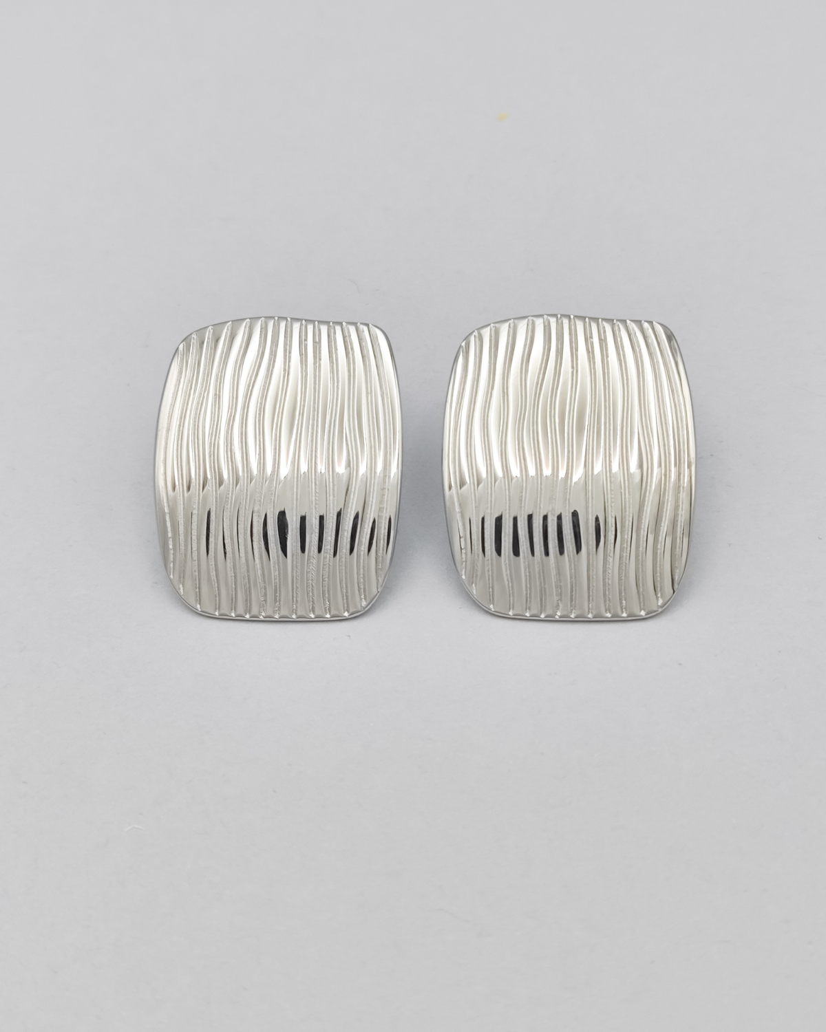 Gli orecchini Marika in acciaio inossidabile anallergico color argento sono rappresentati frontalmente su uno sfondo grigio chiaro. Hanno una forma rettangolare con delle righe verticali leggermente ondulate.