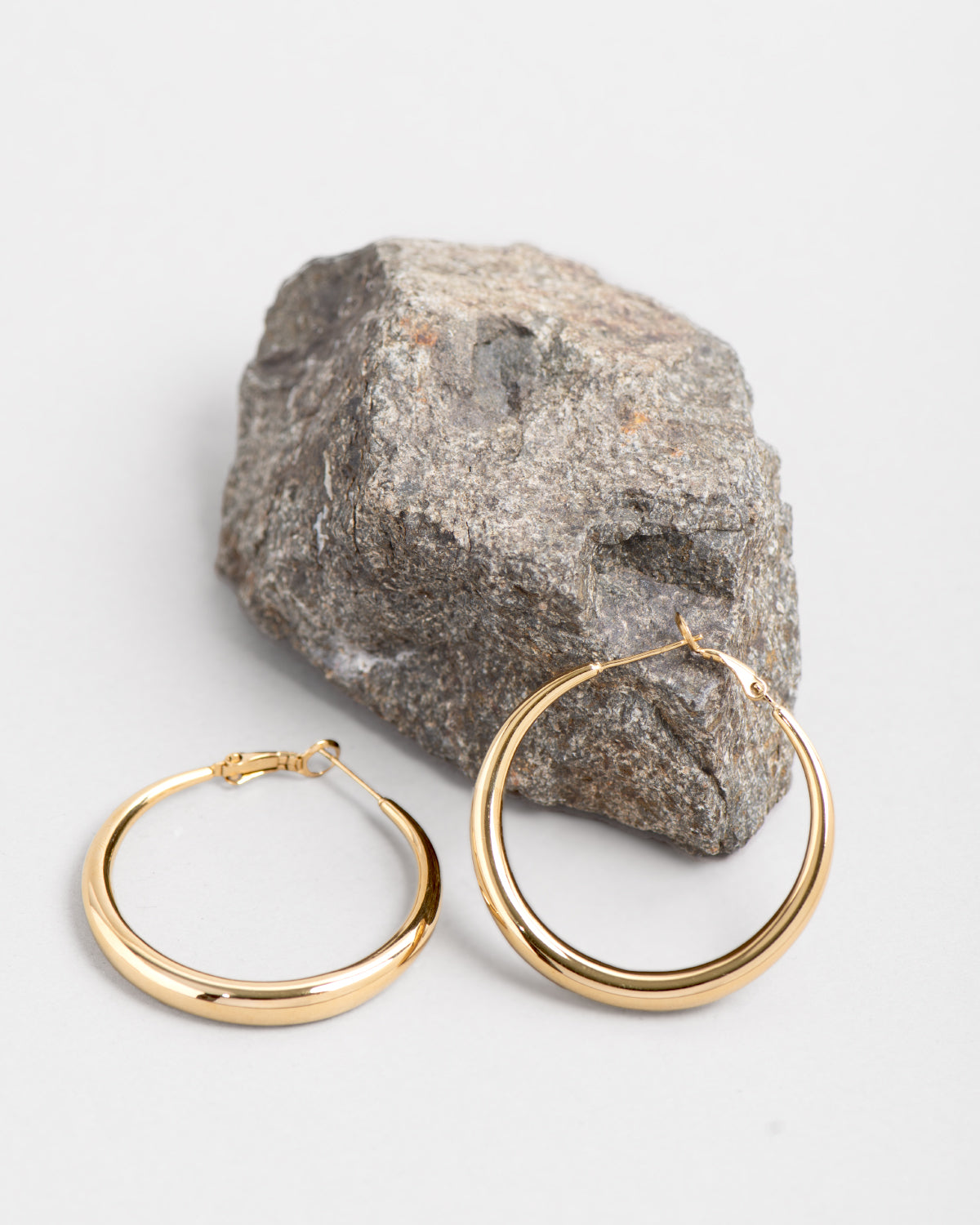 Gli orecchini Martina sono inquadrati con una roccia color grigio scuro su uno sfondo chiaro. Gli orecchini sono dei cerchi più stretti alle estremità e più spessi al centro. Sono in acciaio inossidabile di colore oro con finish lucido.