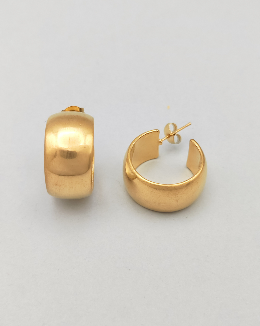 Orecchini Susanna in acciaio inossidabile anallergico color oro. Hanno un design a cerchio, sono molto spessi ma il diametro è contenuto. Hanno un finish opaco. L'orecchino di sinistra è rappresentato frontalmente mentre quello di desta è disteso sul fiano e mostra la chiusura a perno.