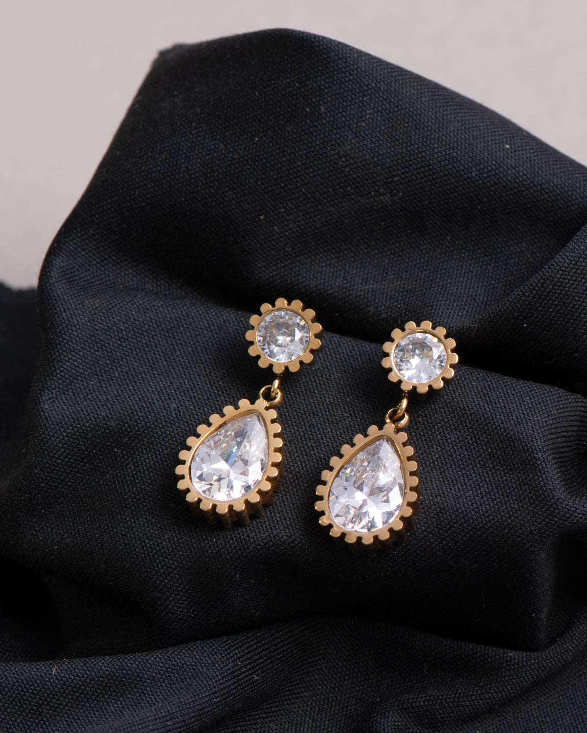 Orecchini Ylenia in acciaio inossidabile color oro. Sono appoggiati su un panno nero. Hanno una forma a bottoncino in strass dal quale pende un ciondolo a forma di goccia, sempre in strass.