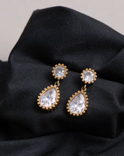 Orecchini Ylenia in acciaio inossidabile color oro. Sono appoggiati su un panno nero. Hanno una forma a bottoncino in strass dal quale pende un ciondolo a forma di goccia, sempre in strass.