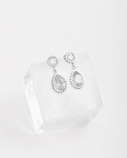 Orecchini Ylenia in acciaio inossidabile color argento. Sono appoggiati su un cubo di cristallo. Hanno una forma a bottoncino in strass dal quale pende un ciondolo a forma di goccia, sempre in strass.