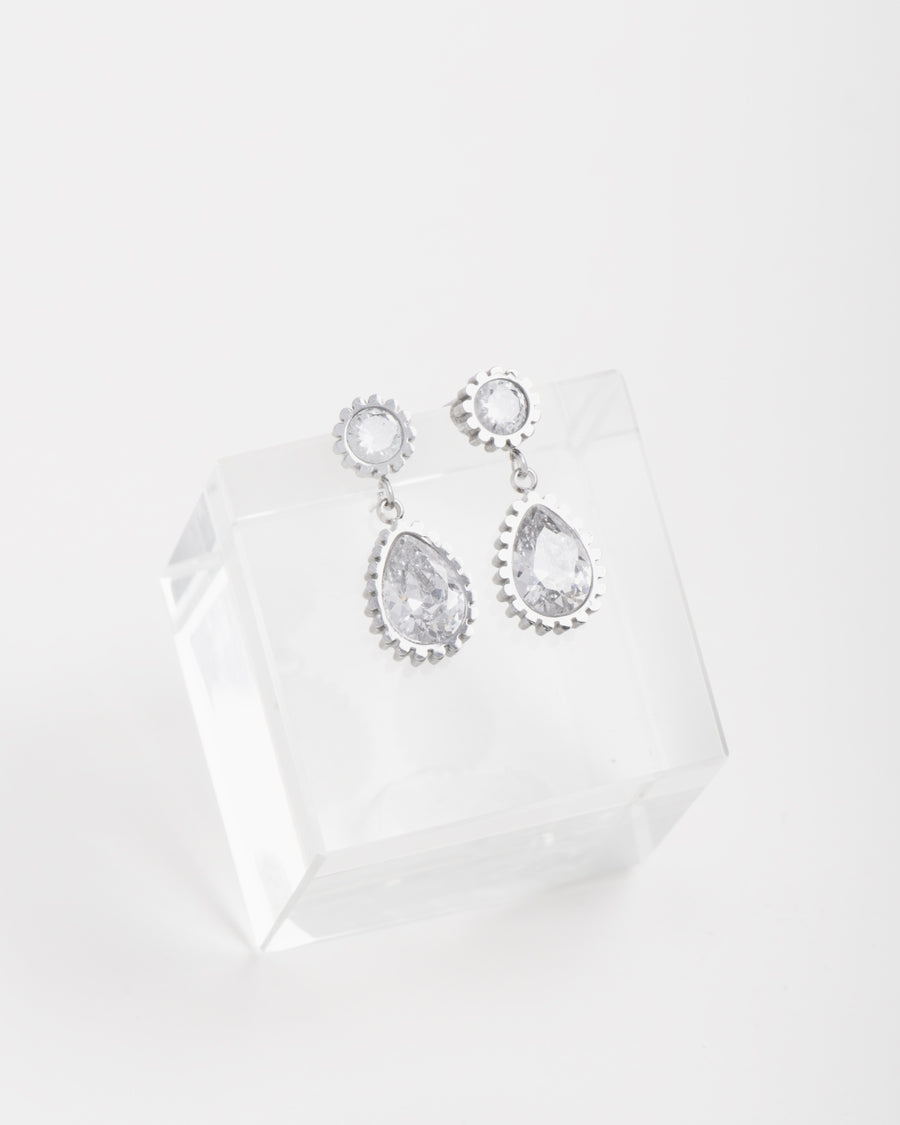 Orecchini Ylenia in acciaio inossidabile color argento. Sono appoggiati su un cubo di cristallo. Hanno una forma a bottoncino in strass dal quale pende un ciondolo a forma di goccia, sempre in strass.
