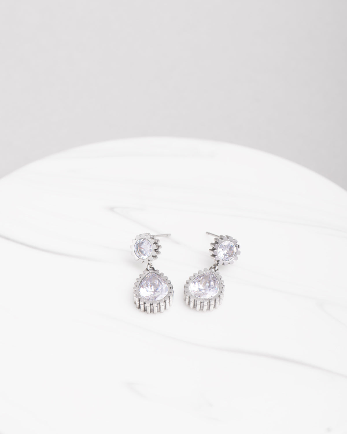 Orecchini Ylenia in acciaio inossidabile color argento e strass. Sono adagiati su una superficie ad effetto marmo chiaro. Dal bottoncino in strass scende un pendente a goccia, anch'esso in strass. Il bordo in acciaio è zigrinato.