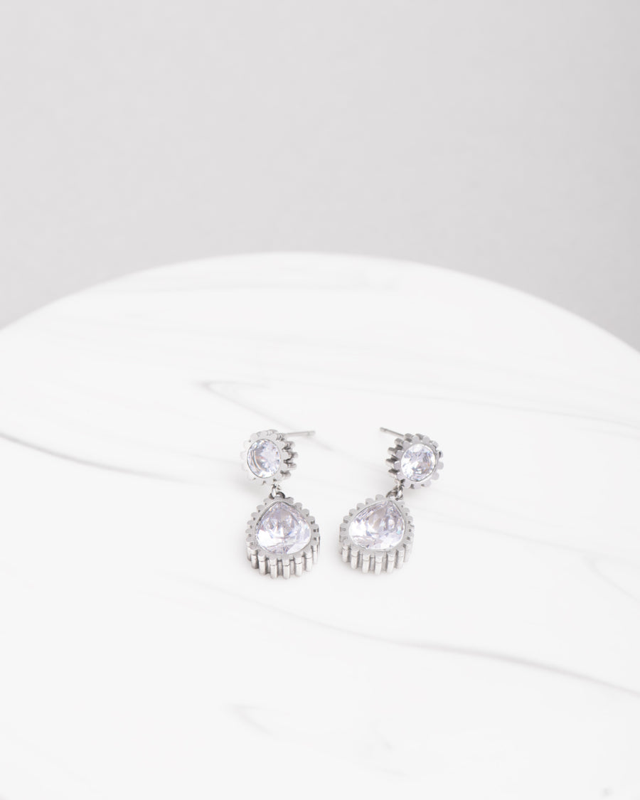 Orecchini Ylenia in acciaio inossidabile color argento e strass. Sono adagiati su una superficie ad effetto marmo chiaro. Dal bottoncino in strass scende un pendente a goccia, anch'esso in strass. Il bordo in acciaio è zigrinato.