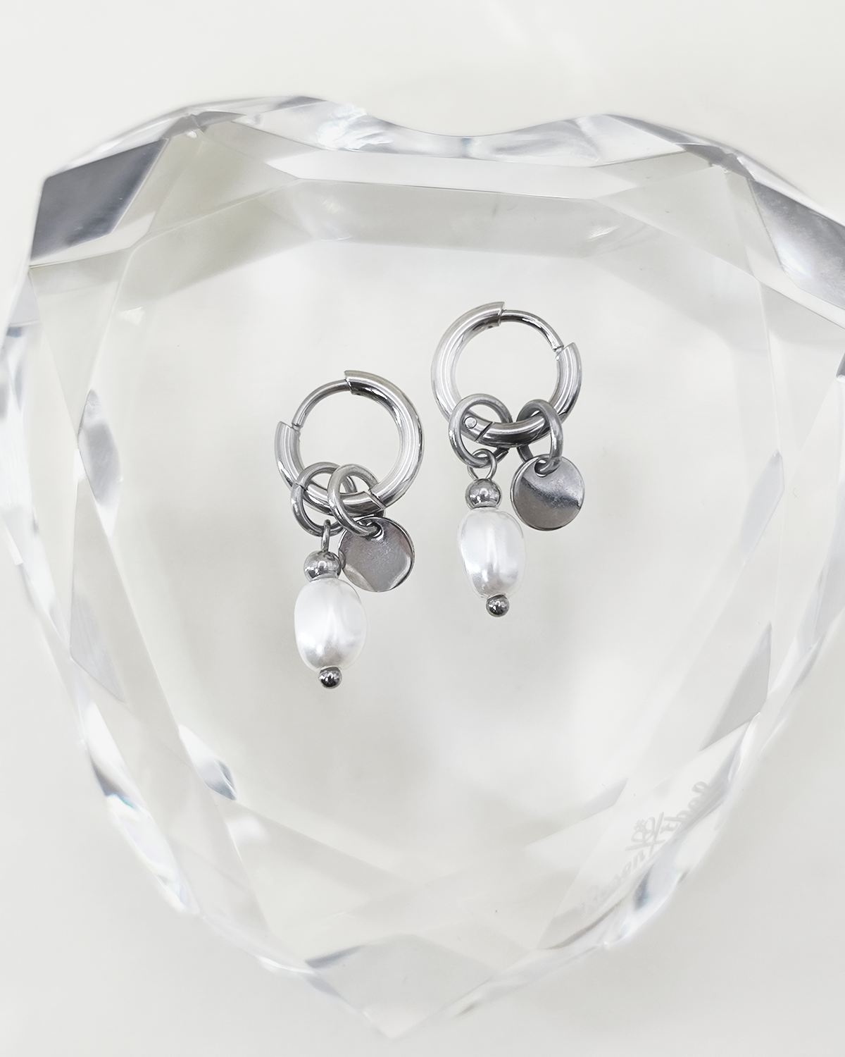 Orecchini Aurora color argento in acciaio inossidabile anallergico e waterproof con ciondolo di perla e medaglietta. Sono appoggiati su un cuore di cristallo trasparente.