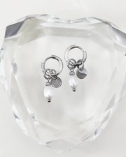 Orecchini Aurora color argento in acciaio inossidabile anallergico e waterproof con ciondolo di perla e medaglietta. Sono appoggiati su un cuore di cristallo trasparente.