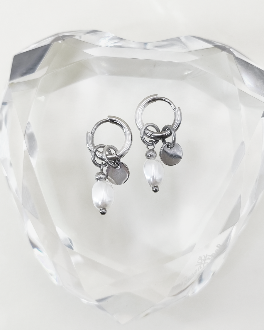 Orecchini Aurora color argento in acciaio inossidabile anallergico e waterproof con ciondolo di perla e medaglietta. Sono appoggiati su un cuore di cristallo trasparente.