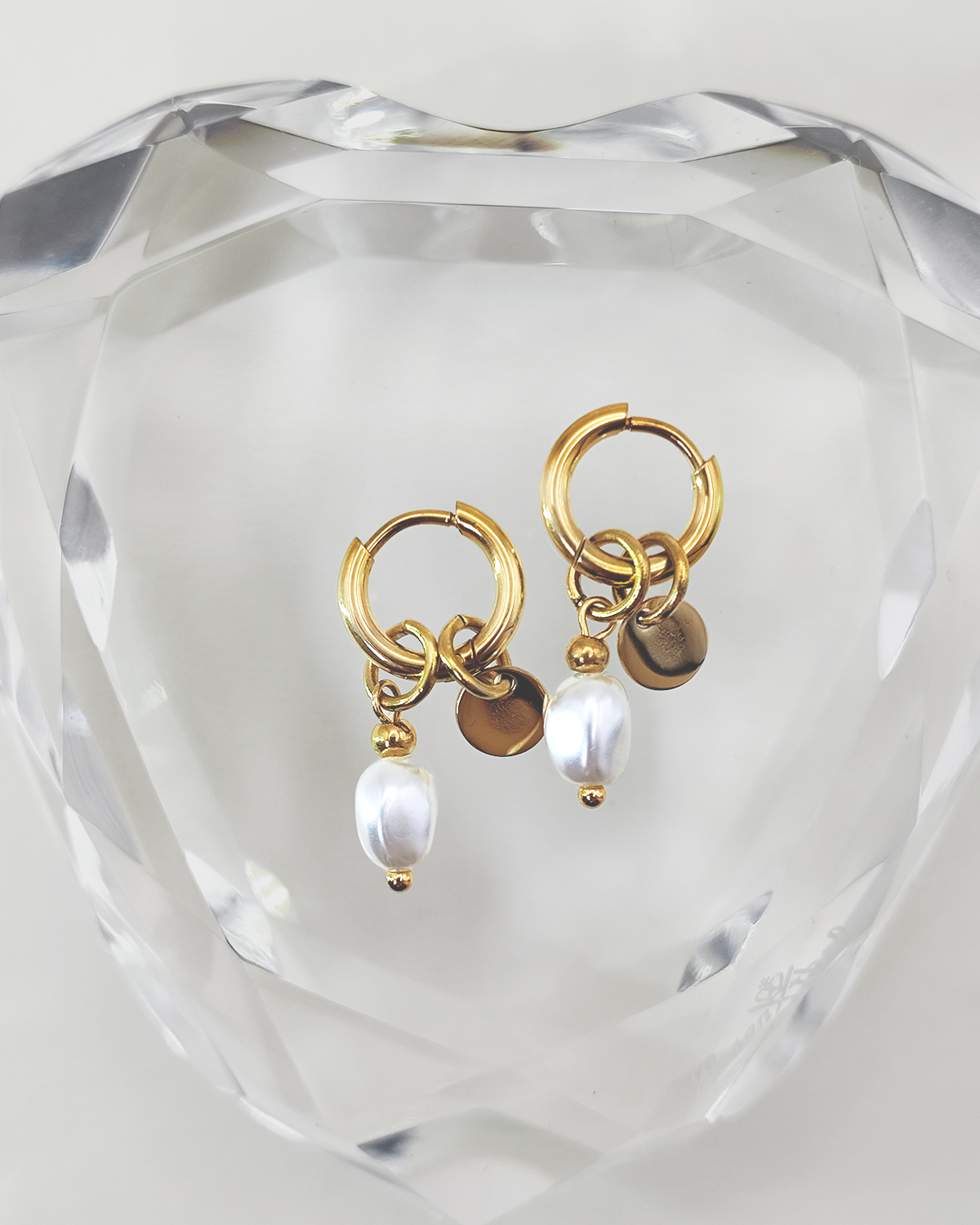 Orecchini Aurora in acciaio inossidabile anallergico e waterproof con ciondolo di perla e medaglietta. Sono appoggiati su un cuore di cristallo trasparente.