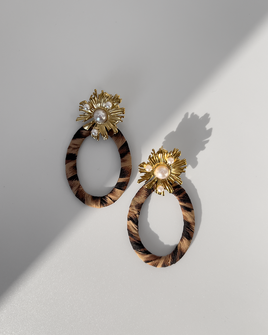 Orecchini Barbara composti da un fiore in acciaio inossidabile color oro e perle dal quale pende un ovale in tessuto maculato. Sono appoggiati su uno sfondo grigio chiaro; l'orecchino di sinistra è in ombra mentre quello di destra è alla luce.