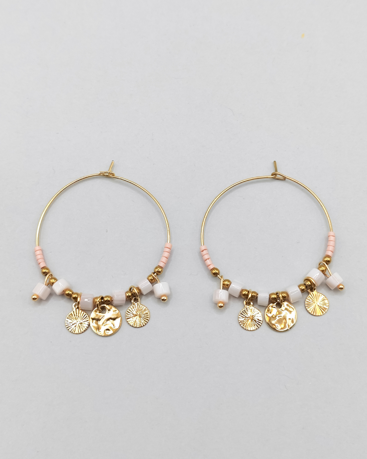 Orecchini Mascia in acciaio inossidabile anallergico color oro. Hanno una forma a cerchio e sono arricchiti da perline e pietre colore rosa e charms dorati.