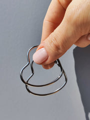 Orecchini Silvia in acciaio inossidabile color argento. Sono sorretti  in modo sfasato da una mano femminile su sfondo grigio. Si nota bene la forma a cuore e il design sottile.