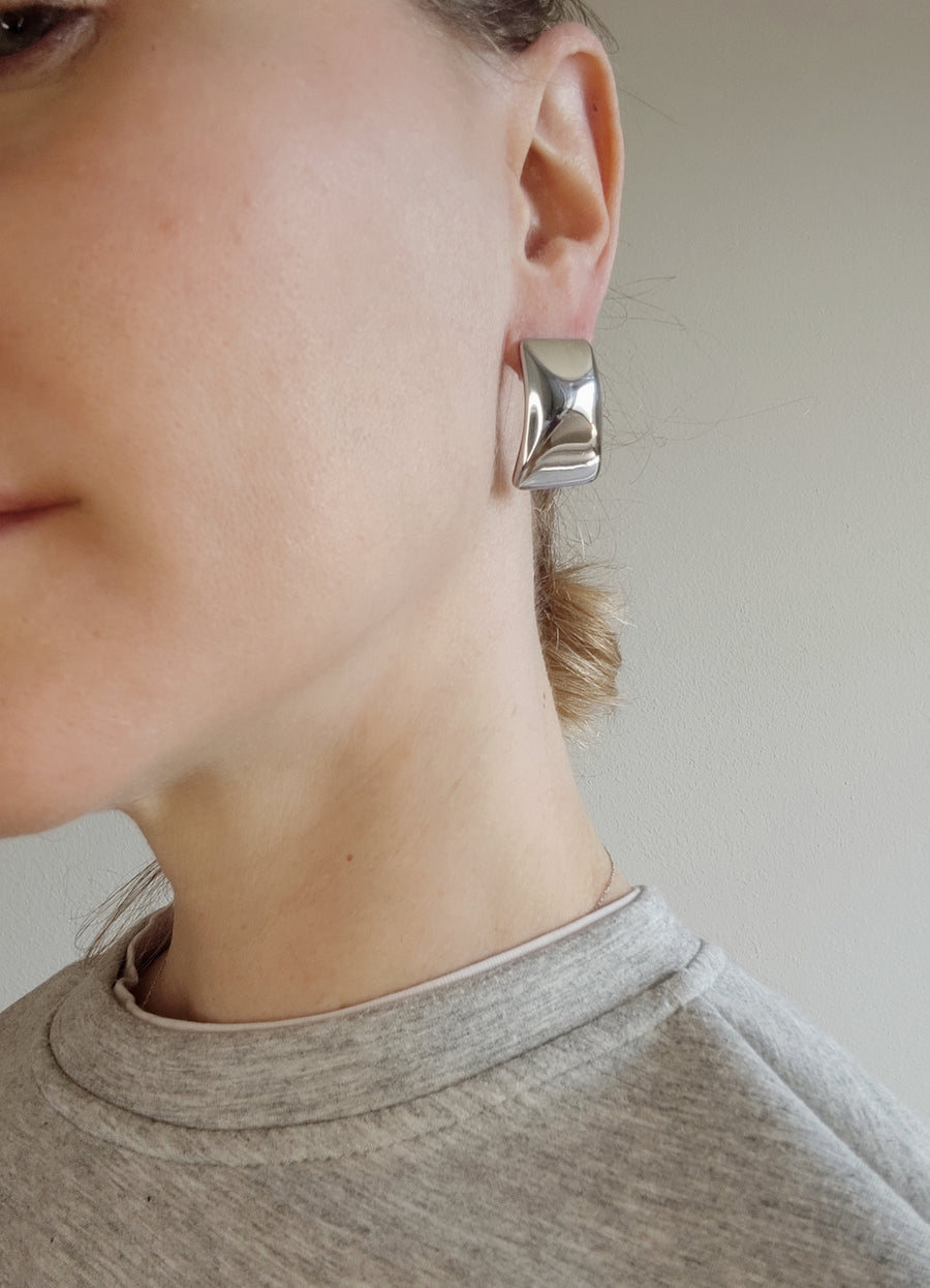 Orecchino Elia in acciaio inossidabile color argento, indossato. La modella veste una tuta grigia che riprende il colore dell'orecchino dalla forma rettangolare, leggermente bombata al centro.