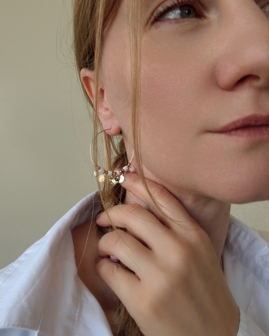 Orecchino a cerchio Mascia in acciaio inossidabile con charms, perle pietre, e cristalli rosa. È indossato da una modella bionda con treccia e capelli biondi.